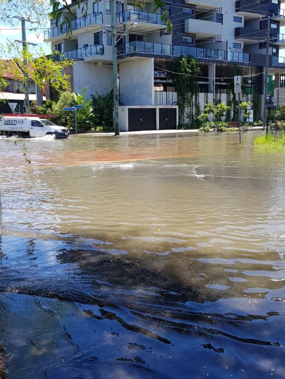 Brown water submerges a road in front of an apartment building. A truck driving in the background has water up to almost the top of its tyres.